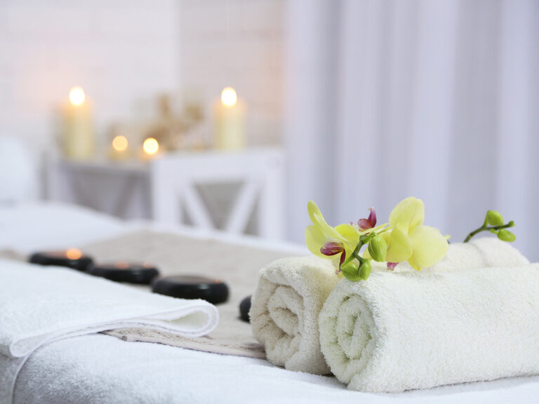 Ein weißes Handtuch liegt auf einer Massagebank, umgeben von Kerzen im Hintergrund.