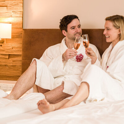 Ein Mann und eine Frau sitzen auf einem Bett und trinken Wein im Hotelzimmer - Landhaus Sponsel-Regus.