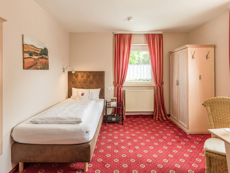 Ein stilvoll dekoriertes Einzelzimmer im Gästehaus, geprägt durch rote Teppichböden und weiße Wände.