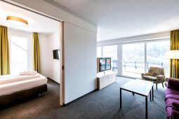 Gepflegtes Hotelzimmer mit einem großzügigen Bett und zwei violetten Sesseln auf Holzboden.